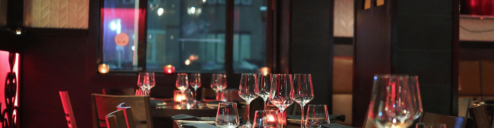 A moody interior shot of a restaurant dining table set with wine glasses and candles, seen through a dark window.