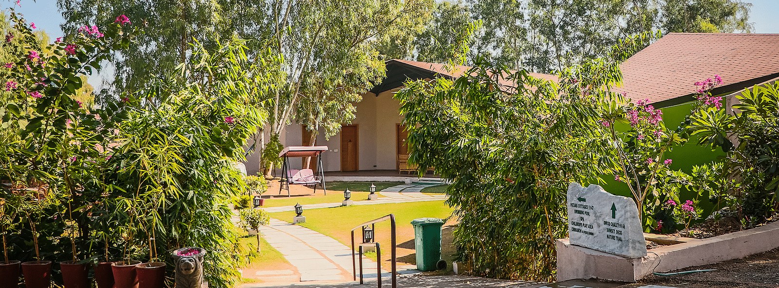 A pathway leads to a building with a sloping roof, surrounded by lush greenery and flowers.- Bamboo Saa Resort & Spa, Udaipur