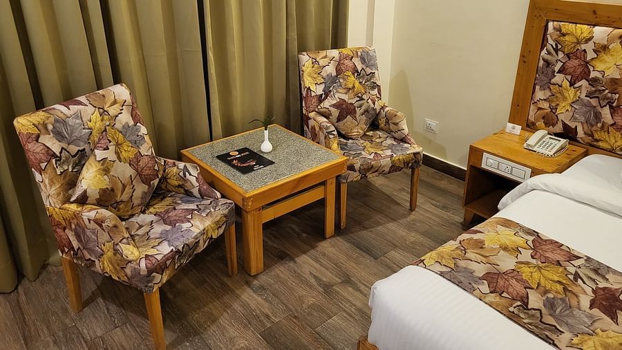 A sitting area with two floral-patterned chairs, a small table, and beige curtains in a hotel room at Dagshai Manor Kasauli - Inde Hotels.