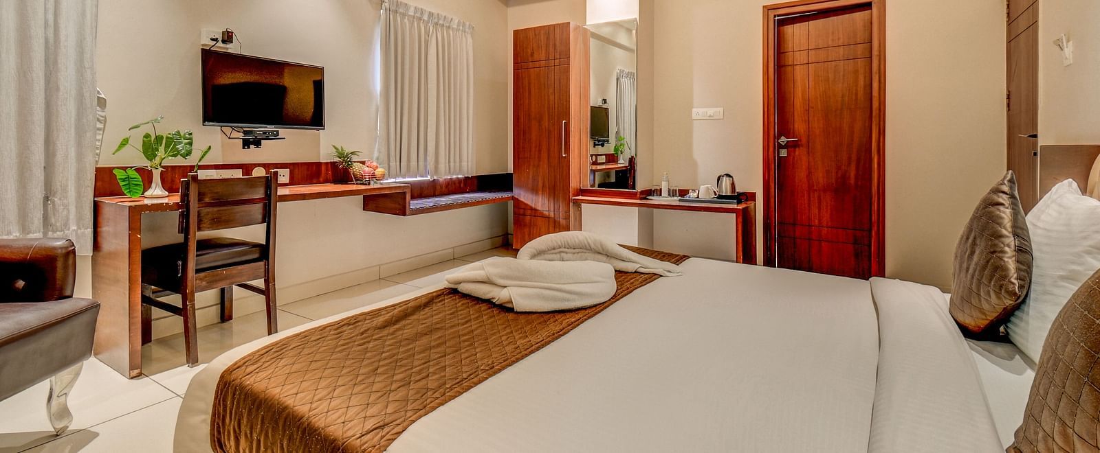 A view of a spacious bed, wall-mounted TV, desk below it, and a chair beside the side table at Harigovinda's JNStay