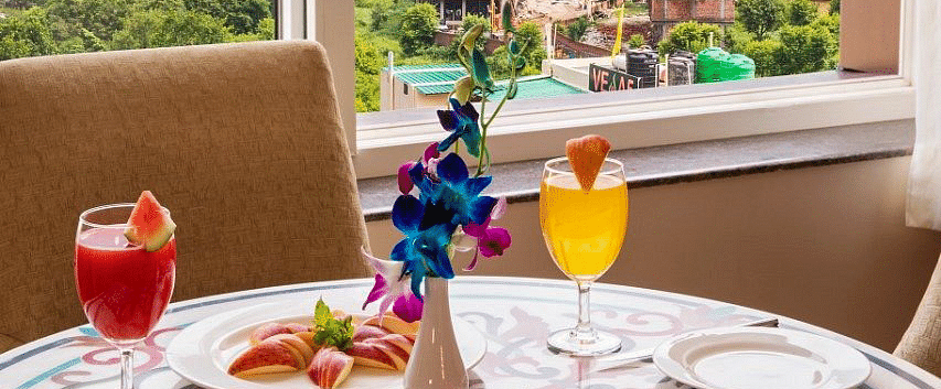 A close up shot of the dining table with well plated fruits and juices overlooking the lush green views from the window at Regenta Inn DLS.