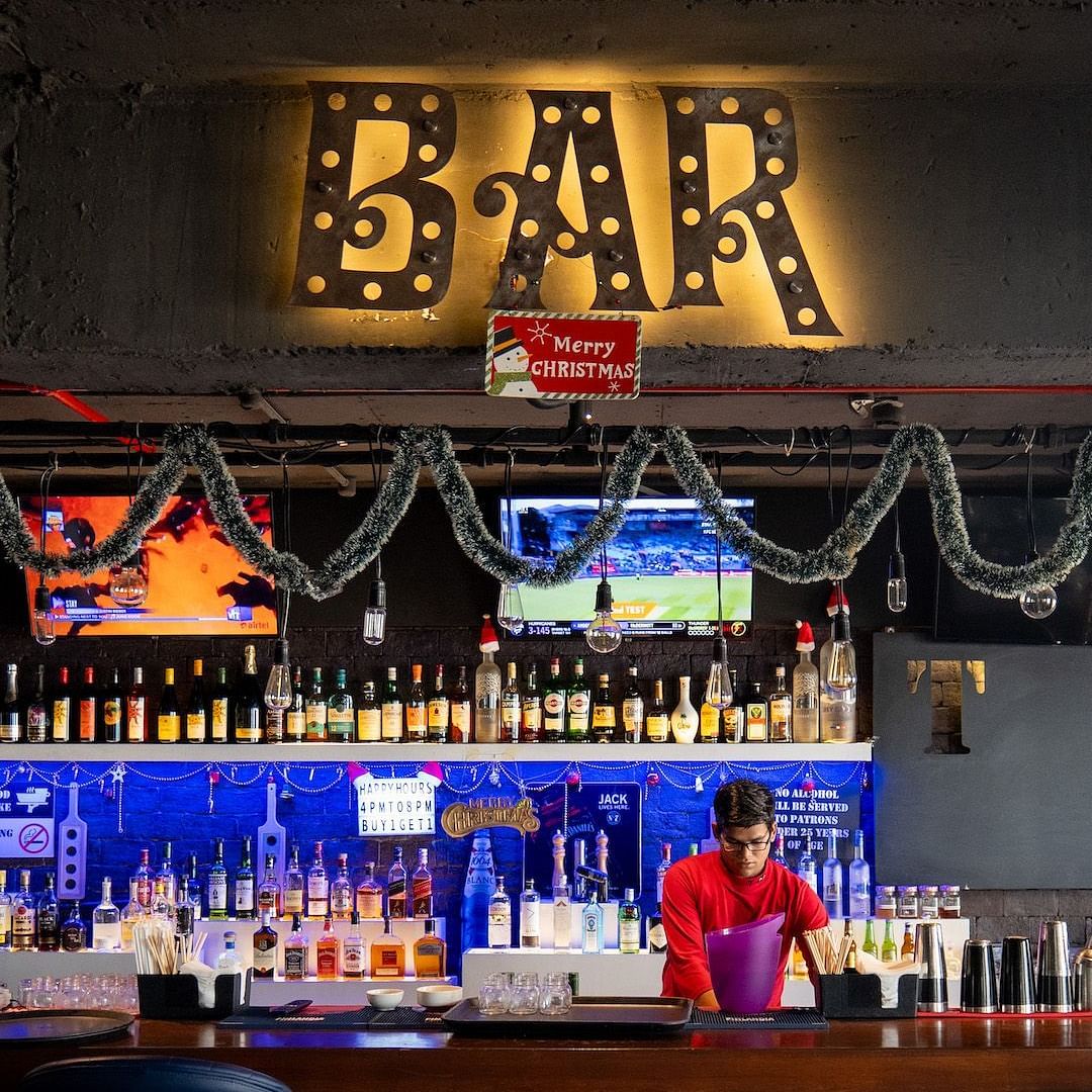 A bartender making a drink behind a brightly lit bar. The bar is decorated for Christmas with lights and garland.
