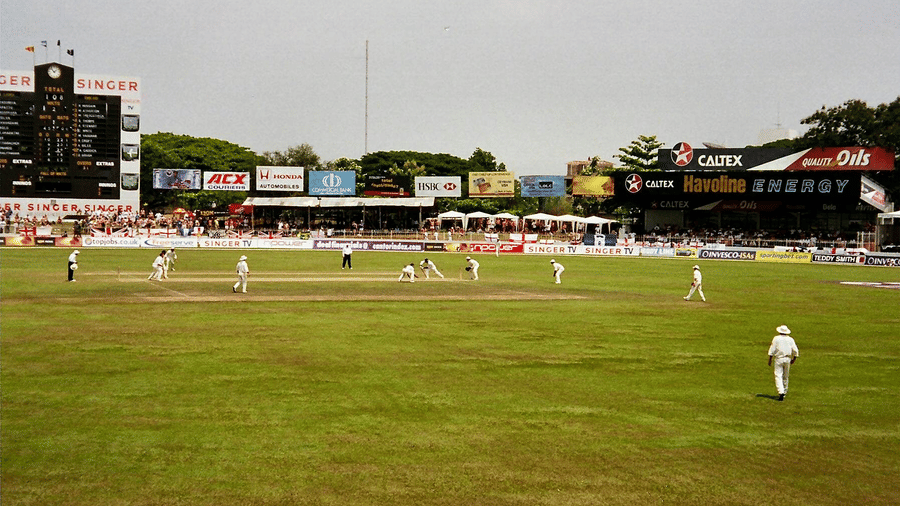 Live cricket match in progress on a smooth green wicket, with fielders spread out and scoreboard buildings in the background.