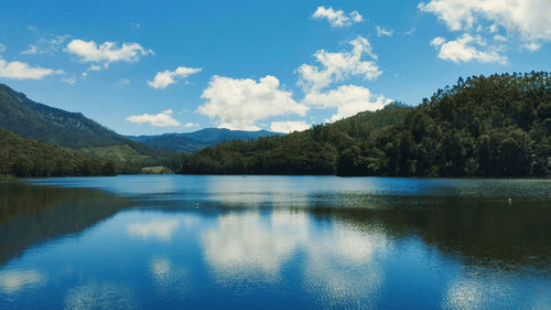 A large, calm blue lake with soft ripples reflecting fluffy white clouds and a bright blue sky, bordered by dense green forests and distant hills.