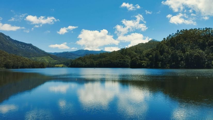 A serene lake reflecting green hills and a bright blue sky with white clouds.