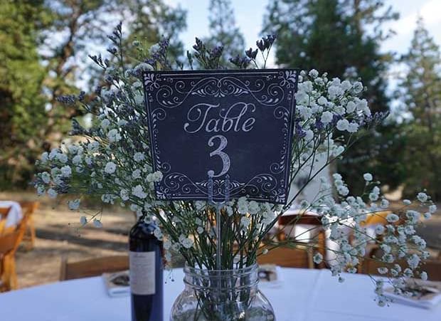 A wedding reception table is set outdoors with a 'Table 3' sign, floral arrangements, and a bottle of wine