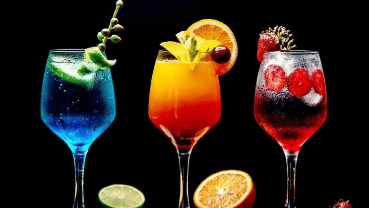 Three colourful fruit mocktails garnished with lime, orange and strawberries served in elegant wine glasses against a black background.