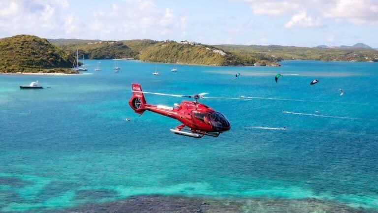 A red helicopter hovers over brilliant turquoise water, with sailboats and kitesurfers visible in the distance and green hills beyond.