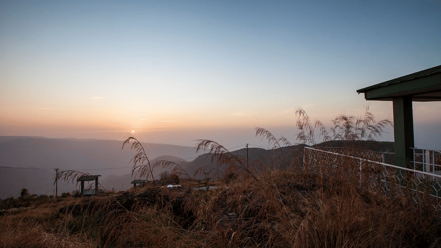 Polo Resort Cherrapunji grounds at sunset with tall dry grasses, low fence, a corner of a green-roofed pavilion and layered hills under a soft sky