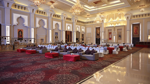Banquet hall at Geeta Sarovar Portico, Panipat featuring table, chairs, and sofa sets in classroom setting. It also buffet counters, chandeliers handing from the ceiling, and huge paintings on the wall.