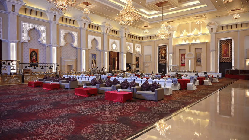 Banquet hall at Geeta Sarovar Portico, Panipat featuring table, chairs, and sofa sets in classroom setting. It also buffet counters, chandeliers handing from the ceiling, and huge paintings on the wall.