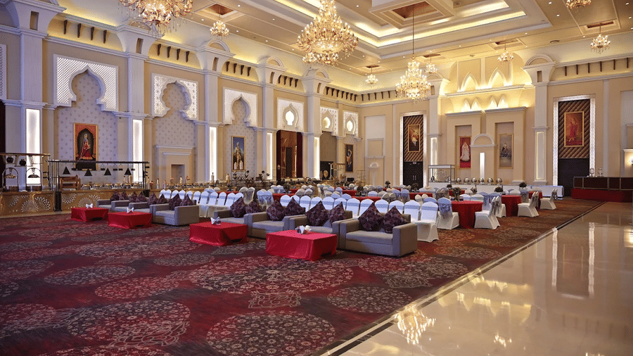 Banquet hall at Geeta Sarovar Portico, Panipat featuring table, chairs, and sofa sets in classroom setting. It also buffet counters, chandeliers handing from the ceiling, and huge paintings on the wall.