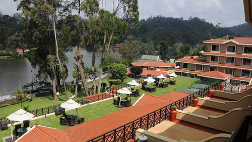 Lakeside view of The Carlton, Kodaikanal, featuring terracotta roofs, manicured lawns, outdoor seating areas, and serene hills reflected in the calm waters.
