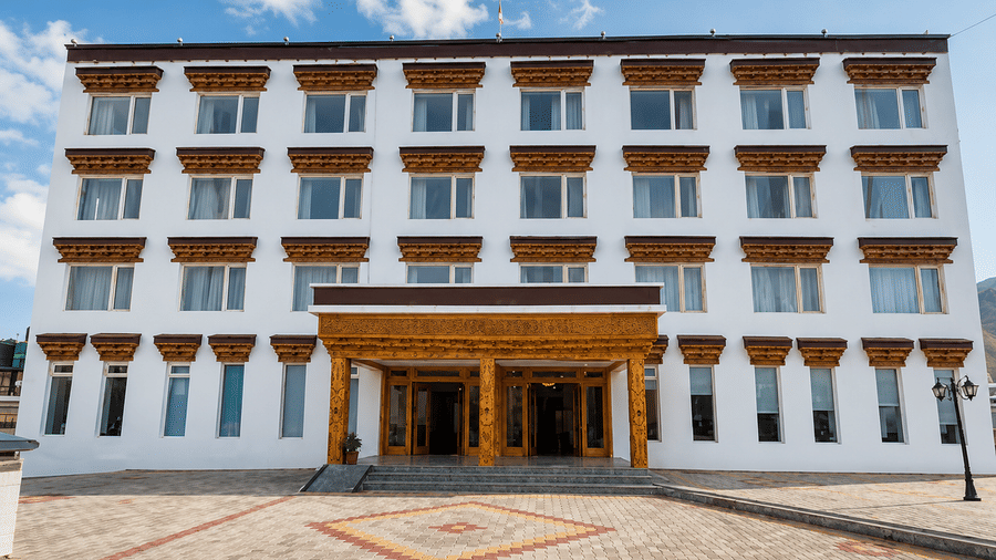 Hotel building exterior in daylight with traditional wooden window frames, a carved entrance canopy and a paved courtyard out front of it.