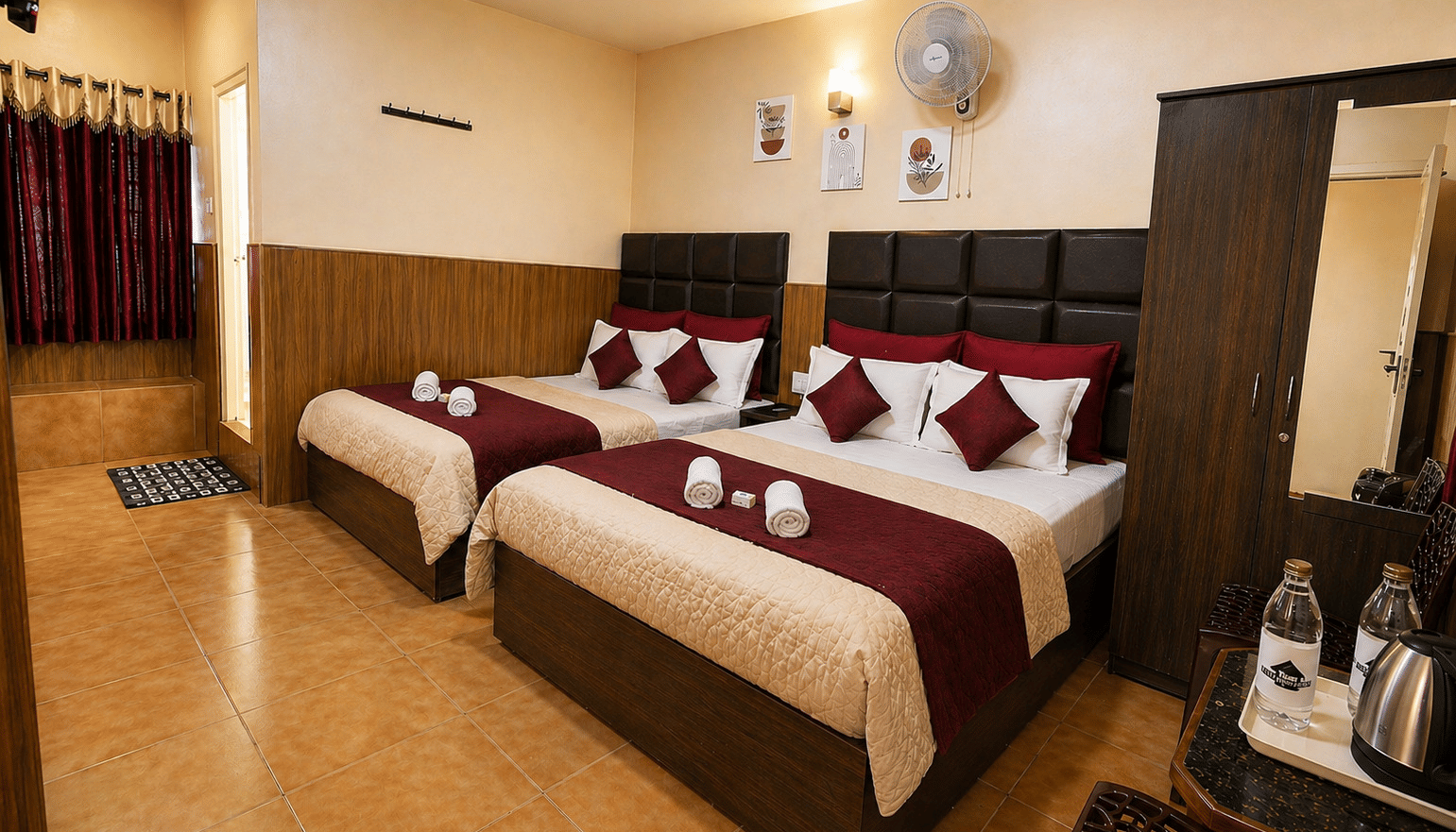 2 large beds arranged side by side with a wardrobe, a wall-mounted fan, and a kettle with water bottles on a table in the Deluxe Quadruple Room at BN Tourist Home, Ooty.