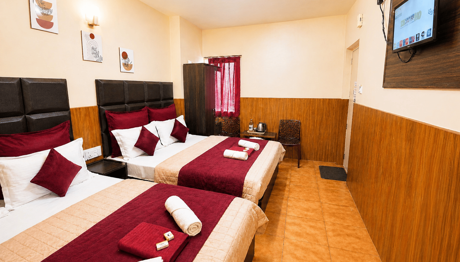 Deluxe Quadruple Room at BN Tourist Home, Ooty, featuring 2 plush beds facing a wall-mounted television screen next to a wardrobe.