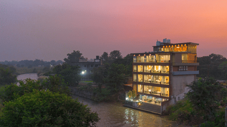 The illuminated Sukoon by the Ganges hotel is captured at sunset, sitting gracefully by the brown-toned river water.