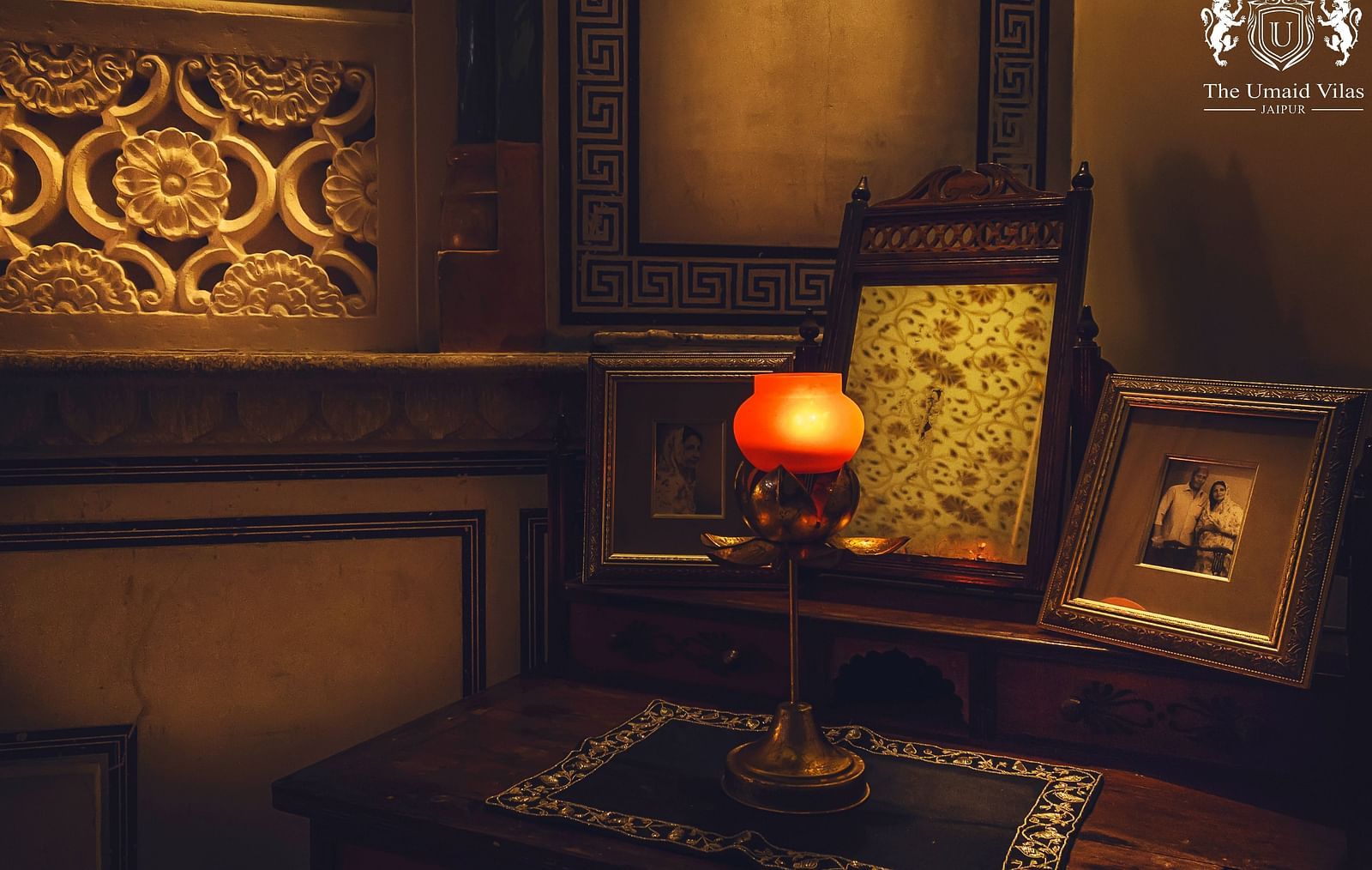 A dimly lit room at Umaid Vilas Jaipur featuring a decorative wall cutout, a lamp with an orange shade, and framed artwork.