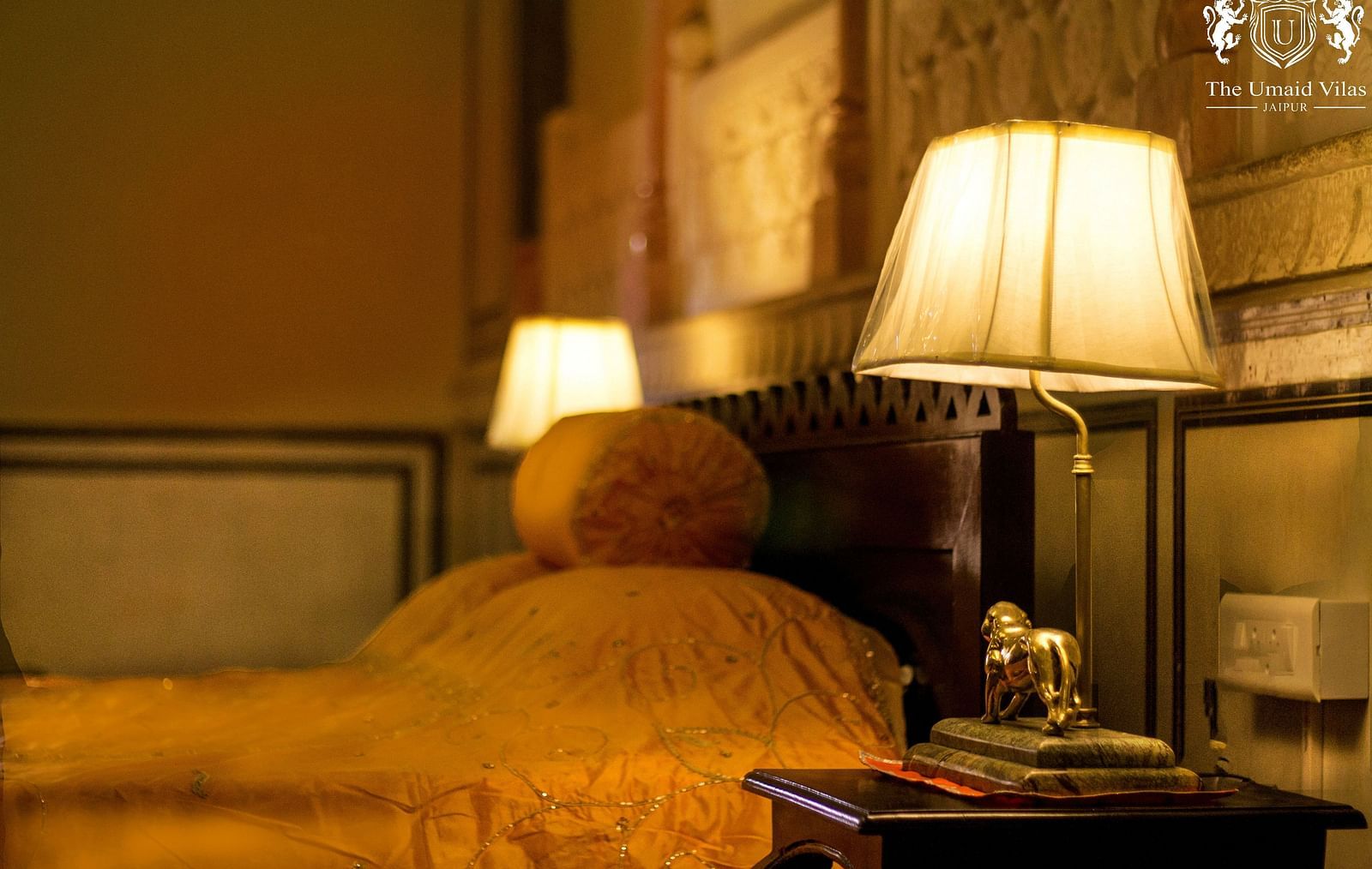 A dimly lit bedroom at Umaid Vilas Jaipur with two lamps on bedside tables, illuminating a bed with a golden throw.
