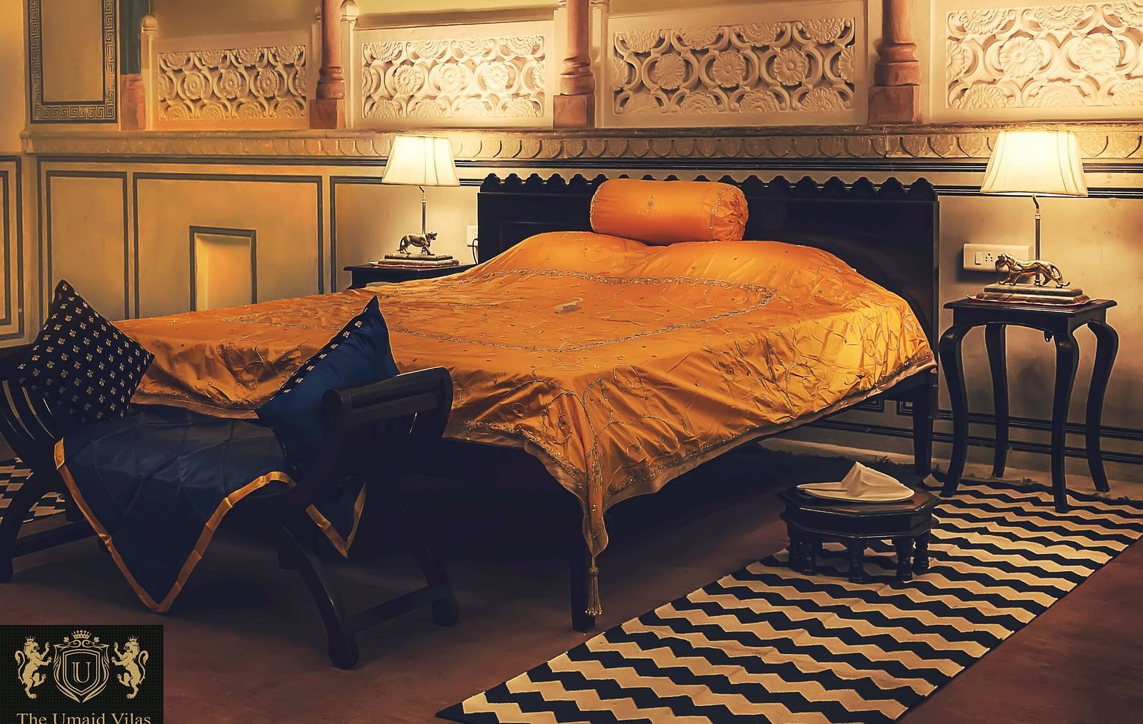 A bedroom at Umaid Vilas Jaipur with a bed dressed in golden fabric, two lamps, and decorative wall cutouts.