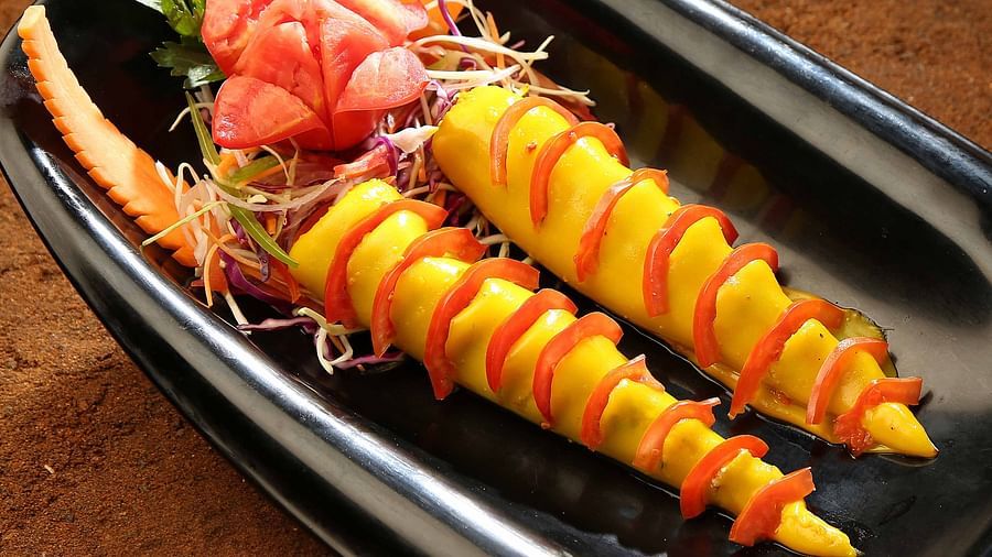Two yellow, spirally cut food items are decorated with red tomato slices and carrot slivers on a black rectangular plate, accompanied by shredded cabbage and parsley - Uday Samudra Leisure Beach Hotel.