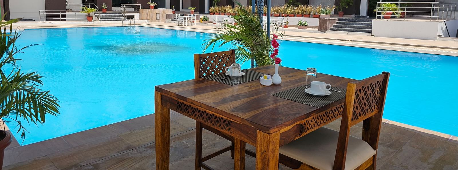 Shot of an outdoor dining area at the Bamboo Saa Mulberry Resort, Pushkar, featuring a wooden table and two chairs set beside a bright blue swimming pool.