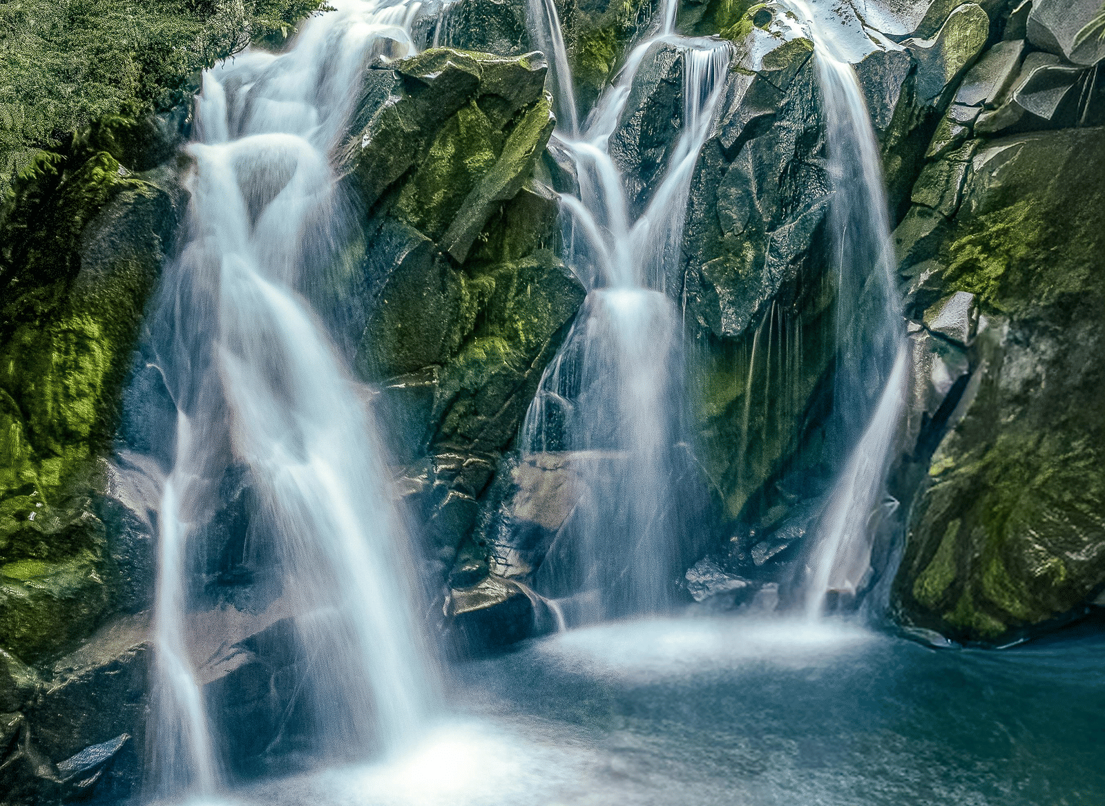 A multi-tiered waterfall cascading over rocky terrain into a pool of water, surrounded by dense green trees