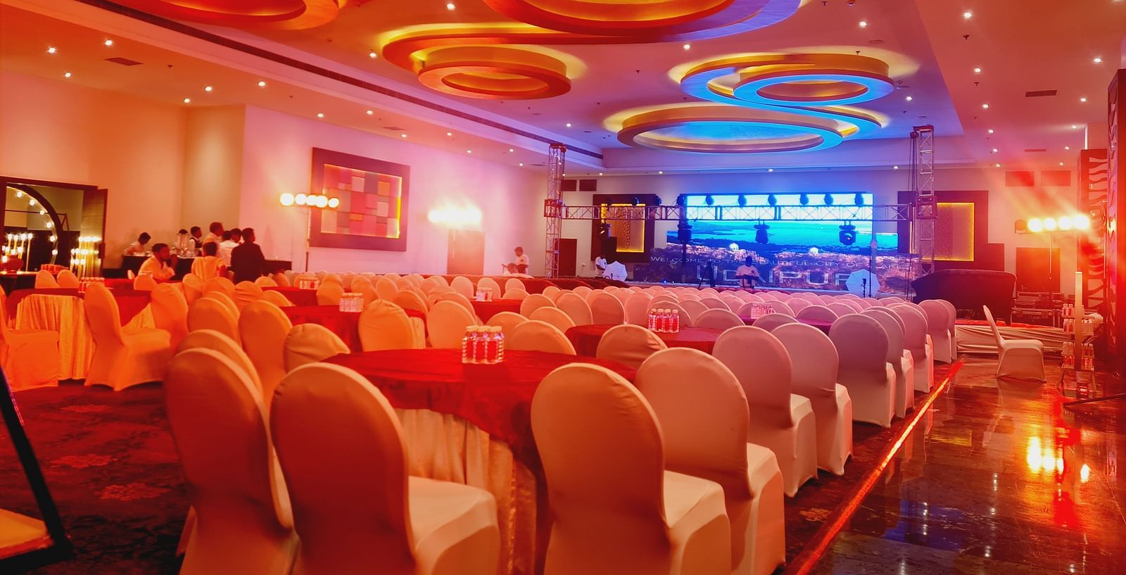 An indoor event space with rows of chairs facing a stage, illuminated by warm lighting and unique circular ceiling fixtures. - Bamboo Saa Resort & Spa, Udaipur