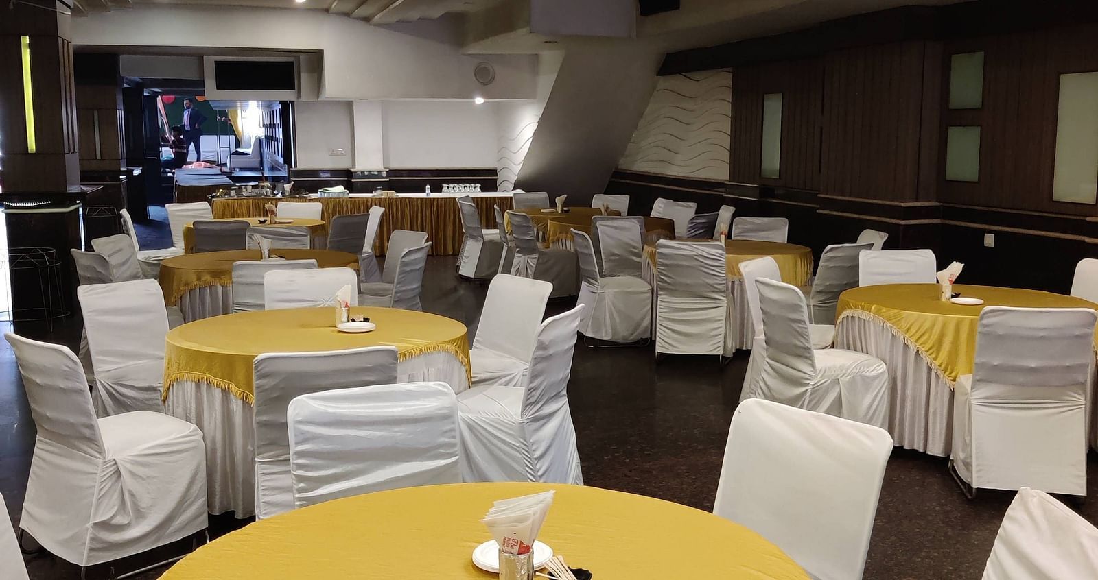 A few round tables covered with bright yellow coloured table cloth and chairs surrounding it with a stage at the far end of the Banquet Hall at Hotel Gold, Panipat.