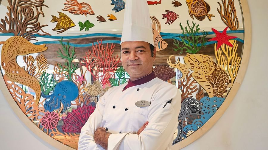 A chef in a white uniform and toque posing with crossed arms in front of a vibrant, artistic wall mural with nature-inspired elements