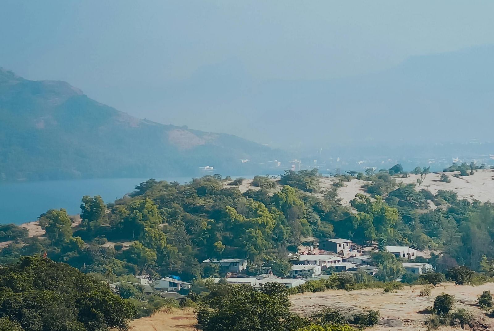 A daytime aerial view of Lonavala, Maharashtra, featuring lush green hills, a small village, and a hazy blue sky.