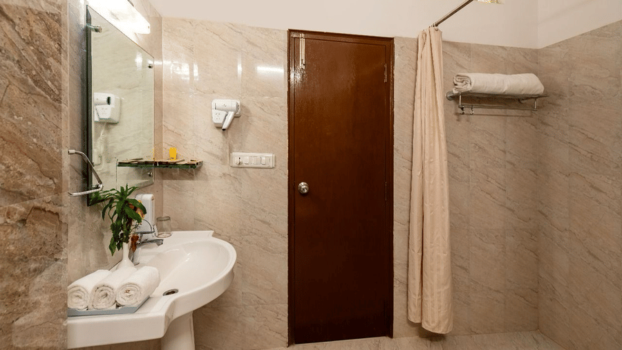 A bathroom showing a pedestal sink with towels, a large mirror, a brown wooden door, and a shower curtain at Amantra Shilpi Resort, Udaipur.