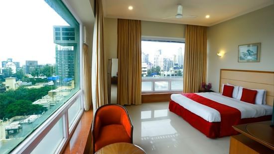 A room adorned with a well-made bed, a coffee table, a red sofa chair, and two large windows offering an expansive city view.