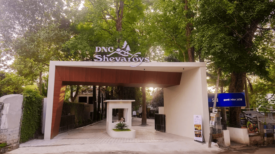 An outdoor entrance shows a gate structure, signage, and trees at DNC Shevaroys Resorts and Spa.