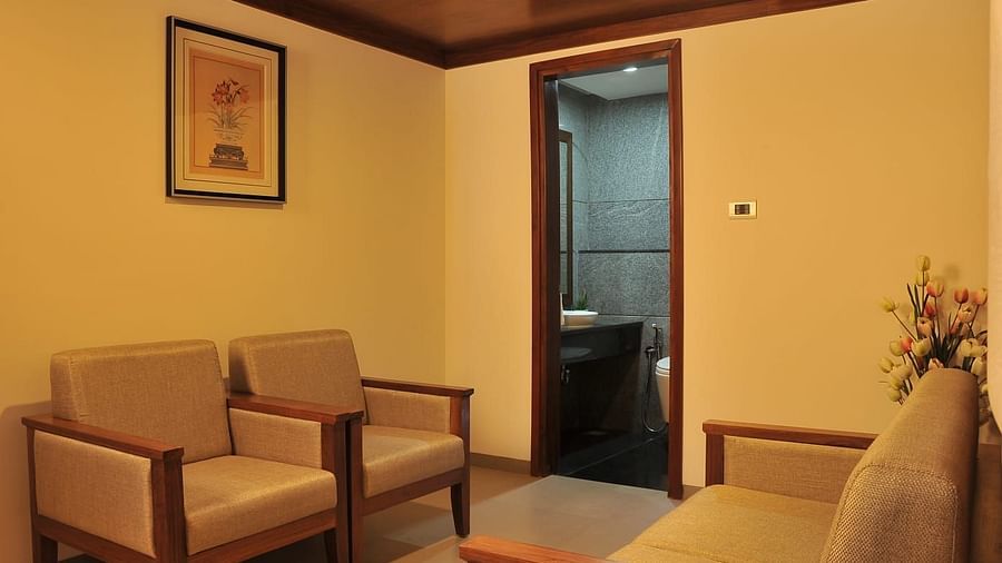 Side view of Suite's seating area at Dhanalakshmi Srinivasan Hotels, Perambalur shows two light brown couches with wooden frames facing each other, a wooden door, and a white vase with artificial tulip flowers.