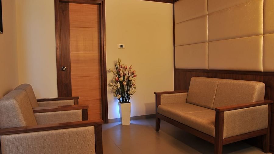 Suite's seating area at Dhanalakshmi Srinivasan Hotels, Perambalur shows two light brown couches with wooden frames facing each other, a wooden door, and a white vase with artificial tulip flowers.