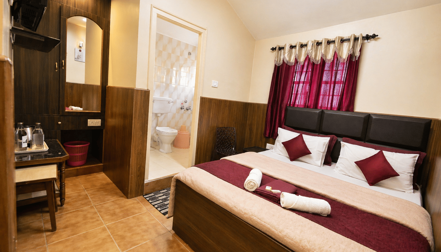 A spacious bed with folded towels, a dressing table with a mirror, and an attached bathroom in the Deluxe Double Room at BN Tourist Home, Ooty.