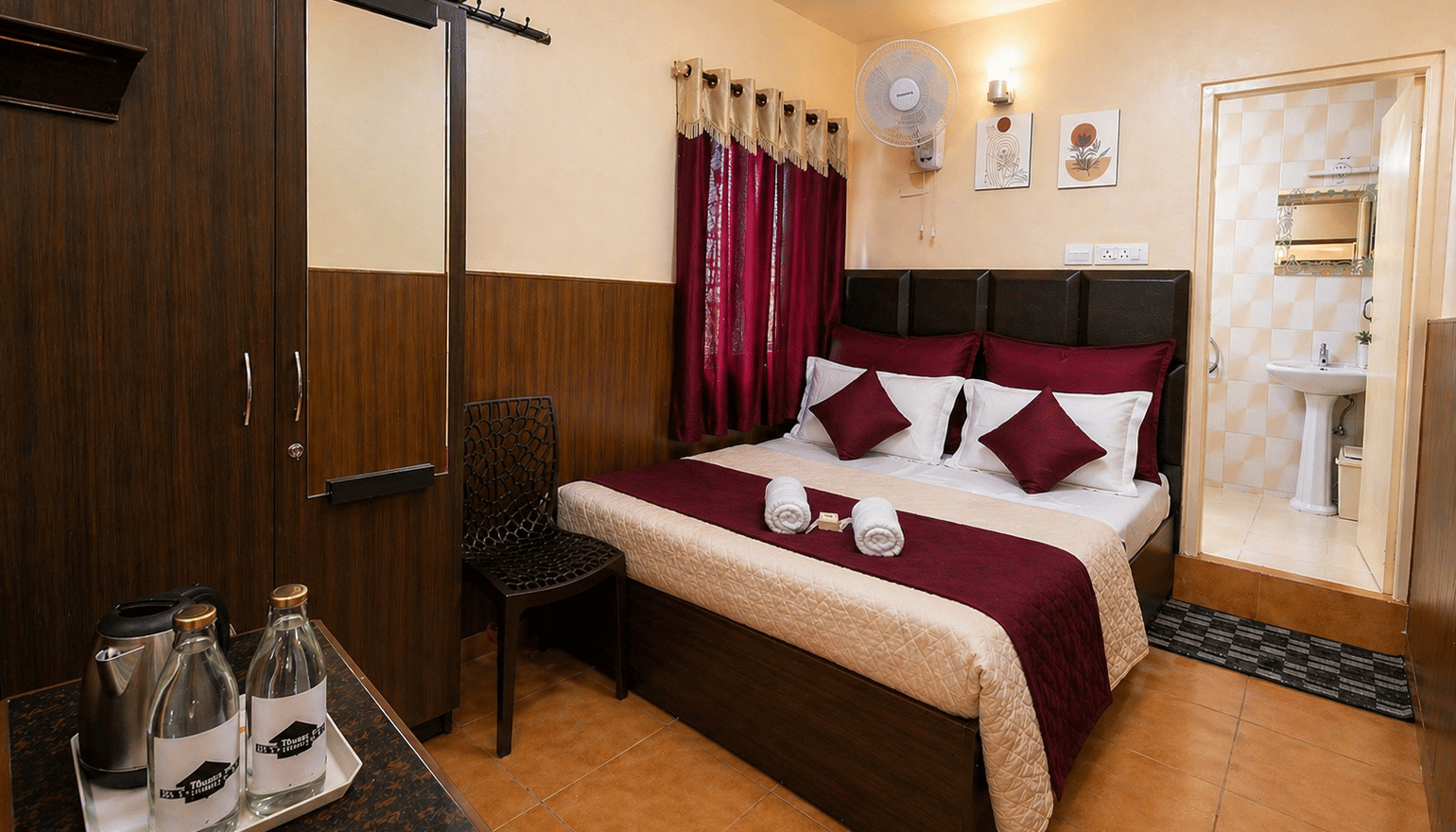 A plush bed next to a large wardrobe with a mirror, a wall-mounted fan, and a kettle setup on a table in the Deluxe Double Room at BN Tourist Home, Ooty.