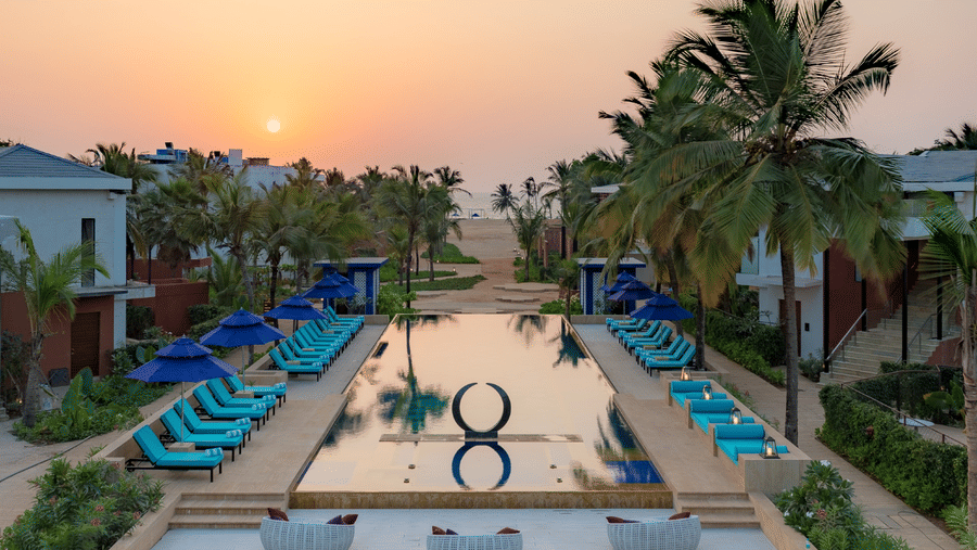 an aerial view of the swimming pool at Azaya Beach Resort, the best stay in South Goa, with loungers and coconut trees lined next to it and the sun setting in the background.