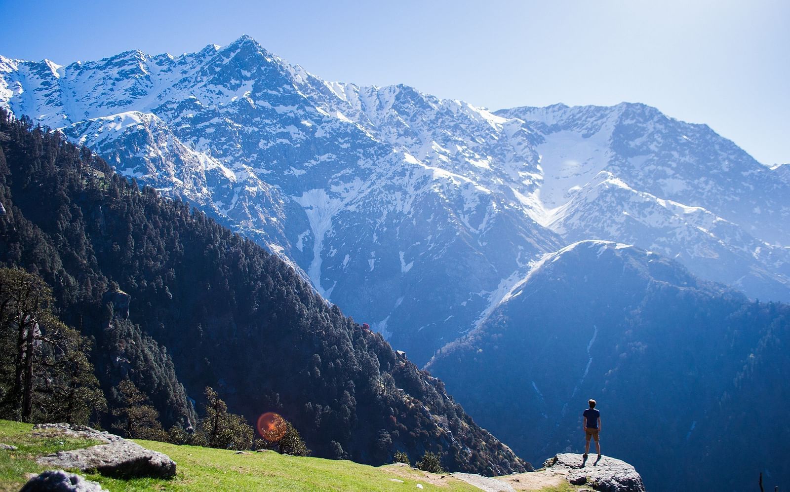 An image of snow caped mountains where a person is seen standing near a cliff enjoying the view