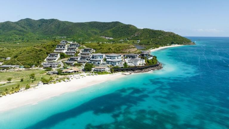 An aerial view of a coastal town with white buildings nestled along a turquoise bay with a white sand beach.