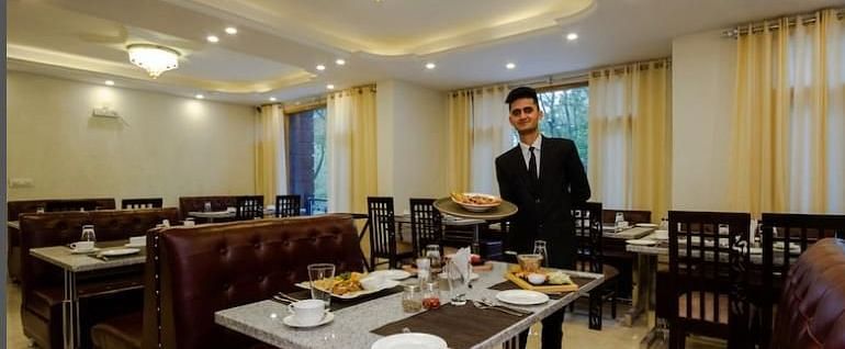 A smiling waiter in a suit holds a tray in a bright dining room with tables set with white tablecloths and silverware | DLS La Serene Manali