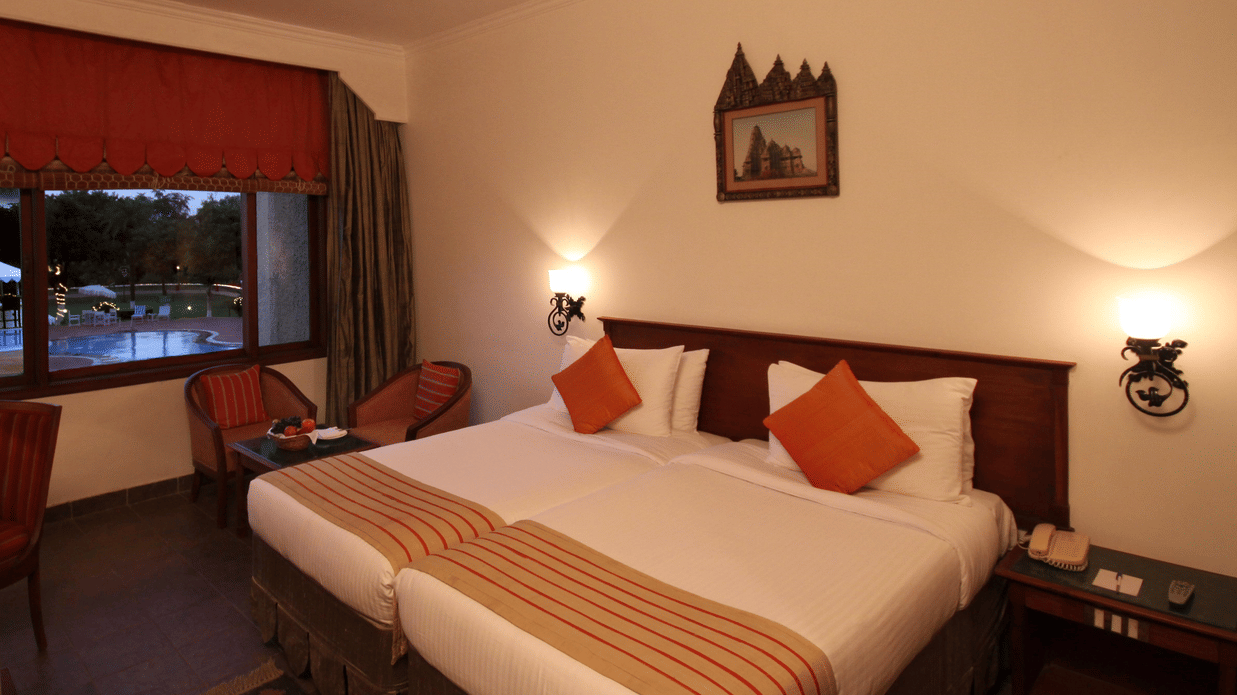 The dusk view of Deluxe Twin Pool View at Clarks Khajuraho, showcasing a large bed and accented pillows featuring dark wood furniture, a ceiling fan, and 2 wall-mounted sconces providing a warm glow. 