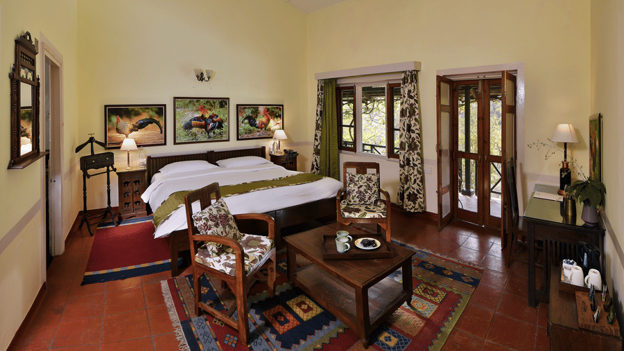 Spacious heritage style suite with a large bed wooden furniture seating area patterned rugs tall ceiling fans and natural light creating a calm and comfortable stay experience at Neemrana’s Glasshouse on The Ganges surrounded by a peaceful indoor atmospher