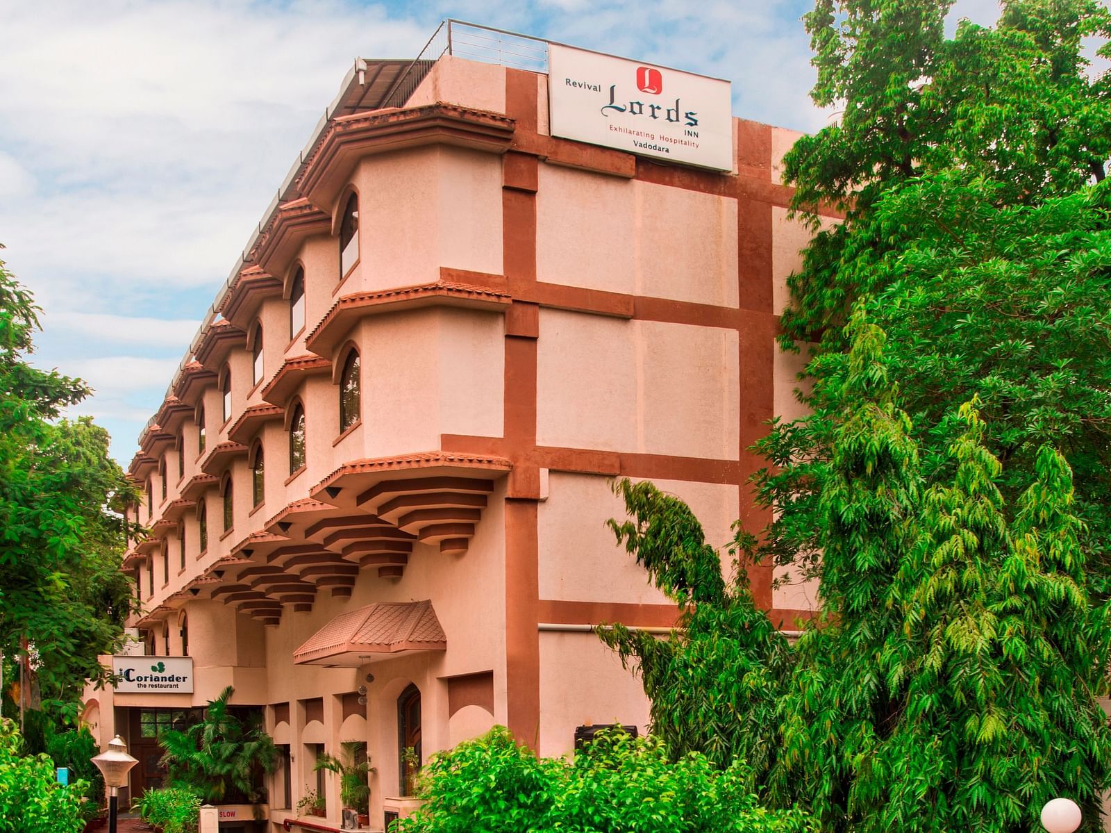 Exterior of Lords Inn Vadodara with a light brown multi-storey hotel surrounded by green trees under a partly cloudy sky.