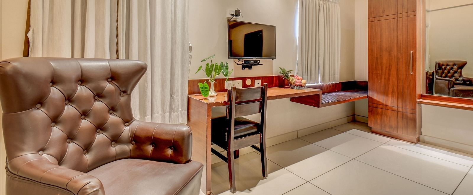 A spacious room at Harigovinda's JNStay with a cushioned chair, wall-mounted TV, desk below it, and a chair