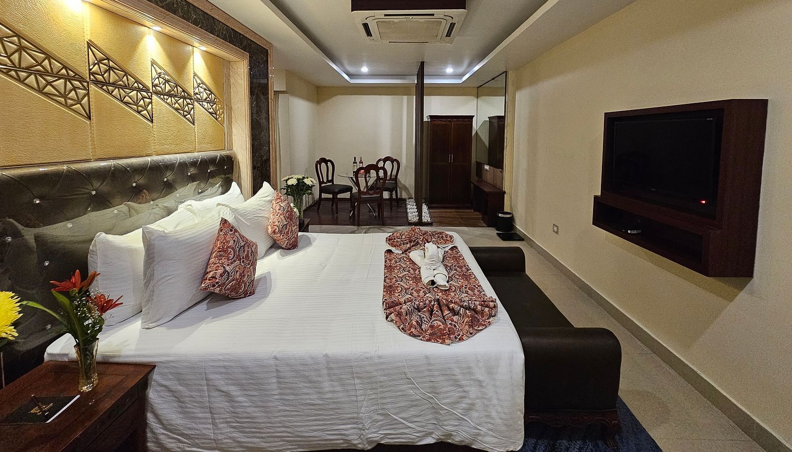 An image of a bedroom with bedside table, a set of table and chairs, flat TV screen, and AC - Sathyam Grand Resort