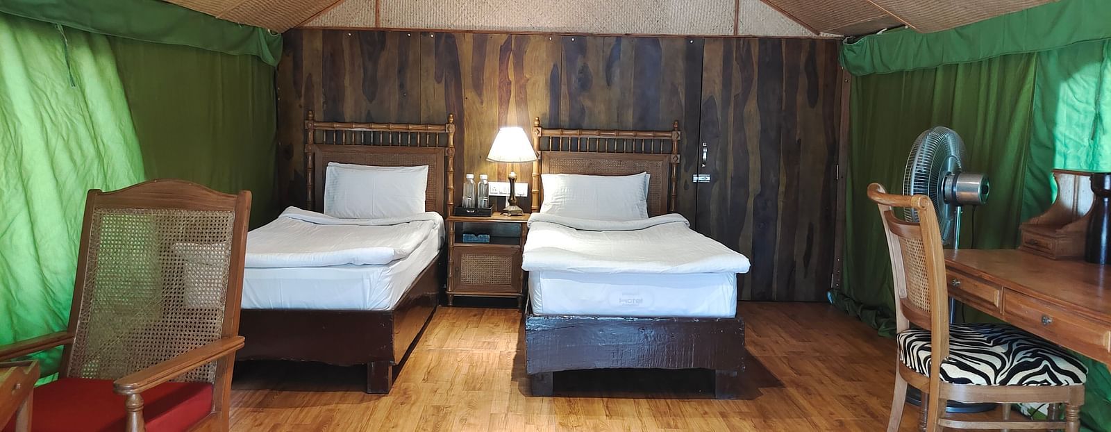 Image featuring Swiss Cottage Tent of The jamoon corbett, with two beds with bed lamp, wooden flooring and a table with two chairs in it.