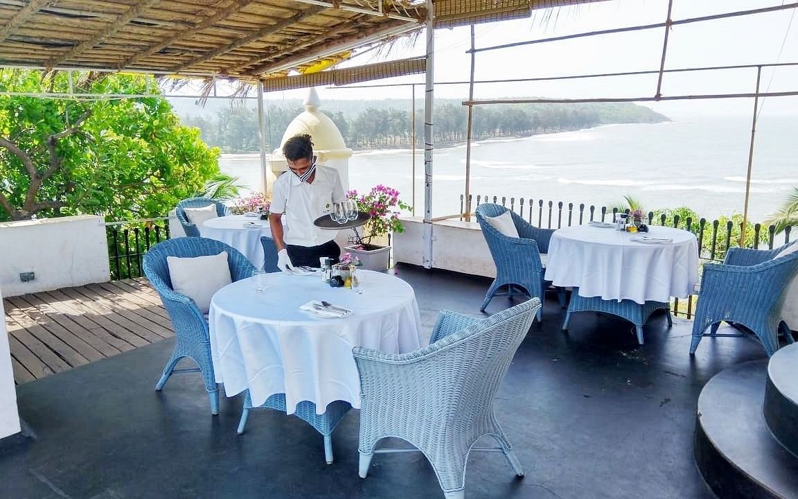 An outdoor dining area at Fort Tiracol, Goa, with tables set with white tablecloths and light blue chairs, under a shaded canopy.