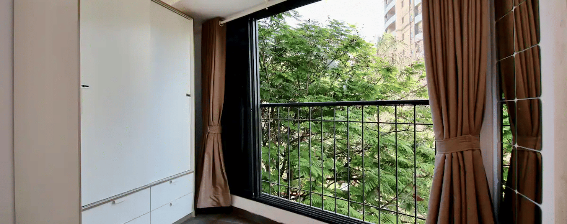 Balcony corner at Wabi-Sabi, Juhu with a white wardrobe, brown curtains, and a black grille window overlooking dense green treetops.